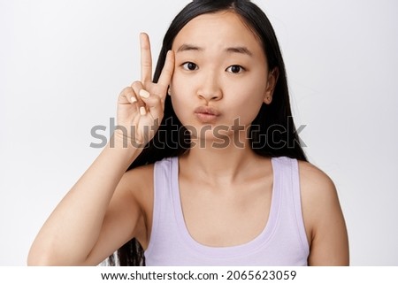 Cute asian woman showing kawaii peace sign near clean face without makeup, standing over white background