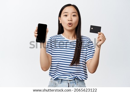 Portrait of asian girl looks surprised while shows empty phone screen and credit card, recommending smartphone application, standing over white background