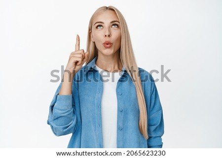 Surprised blond woman pointing and looking up, pucker lips intrigued by something interesting above, standing against white background