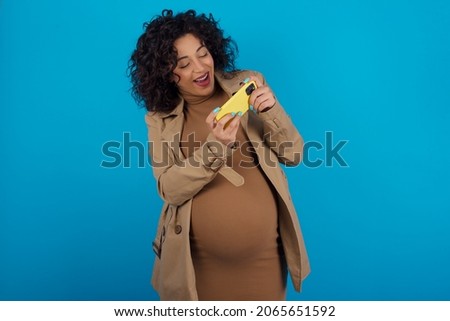 Young pregnant arab woman standing on blue studio background  holding in hands cell playing video games or chatting