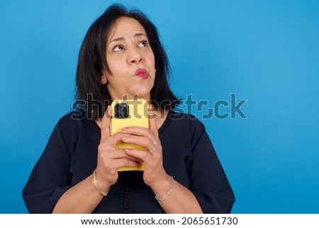 Portrait of middle aged Arab woman standing against blue background with dreamy look, thinking while holding smartphone. Tries to write up a message.