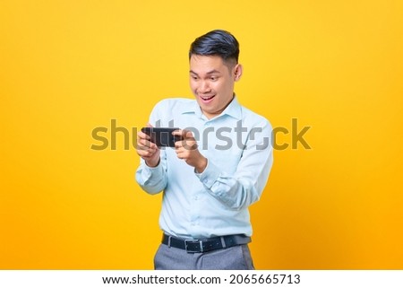 Excited young handsome businessman playing a game on mobile phone on yellow background