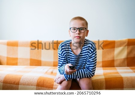 A teenage boy watches tv, using remote control to change channels. Young man is unhappy with what he sees on the tv screen. Sitting on the sofa in the living room at home in casual clothes
