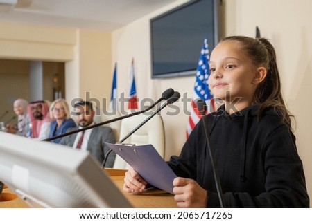 Content confident little eco activist in black sweater standing at rostrum and reading report at political conference Royalty-Free Stock Photo #2065717265