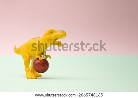 Yellow toy dinosaur tyrannosaurus rex with new year Christmas red baulbe around nack with golden rope on a pink and mint pastel background.