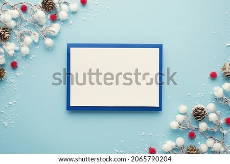 Top view photo of blue envelope with white card christmas decorations snow branches red berries and pine cones on isolated pastel blue background with blank space