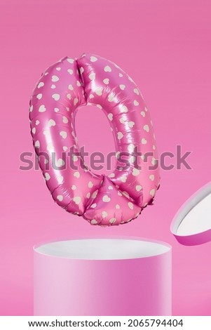 Shot of a pink balloon with white hearts. The balloon has a shape of number 0. The balloon is flying out of the pink box. The balloon is on the pink background.
