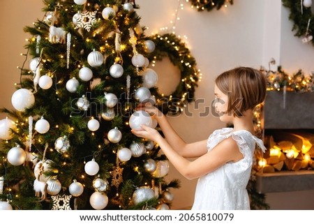 A girl in a white dress decorates the Christmas tree before Christmas at home. Happy childhood. Christmas tree decoration before Christmas.