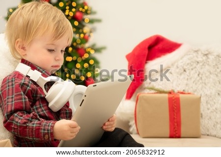 Caucasian baby girl or boy of 1-2 year with headphones looking at tablet indoors.Concept of kid watching cartoons,Christmas film,listen to New Year music,podcast.Toddler with gadget on winter holiday.
