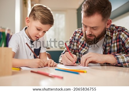 Bearded man in checkered shirt and cute boy drawing pictures with colored pencils while spending time at home together.