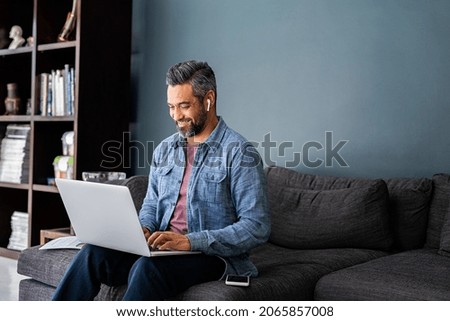 Successful mature indian businessman sitting on couch typing on laptop with wireless earphones. Mixed race businessman sitting on couch while working from home during video call. Business man working  Royalty-Free Stock Photo #2065857008