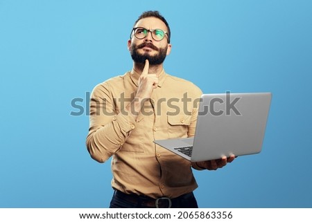 Thoughtful young bearded man holding laptop computer and touching chin with index finger and looking away, isolated over blue background  Royalty-Free Stock Photo #2065863356