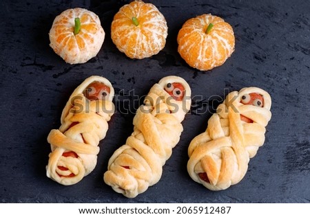 Halloween Egyption Mummy From Sausage And Pastry And Mandarine Shaped Pumpkin On Black Background. Flat Lay.