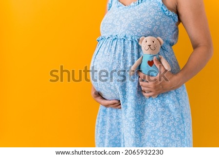 Motherhood, femininity, hot summer - croped portrait pregnant unrecognizable woman in blue dress holds soft toy teddy bear stroking belly expecting baby on yellow bright color background copy space