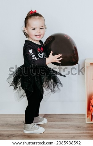 smiling beautiful one year old caucasian girl in halloween costume at home with Halloween decoration, holding balloon. Lifestyle indoors. Halloween party concept.