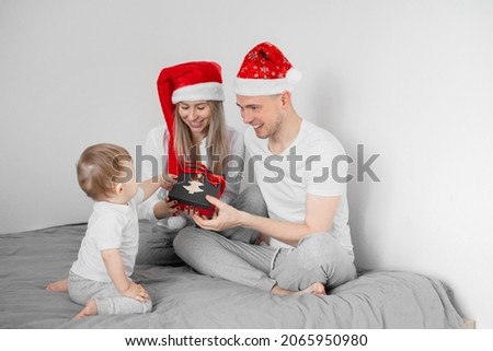 Happy family. Mother and father wearing Santa hats and their baby son opening Christmas presents sitting on bed. White background.