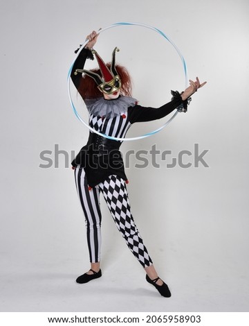 Full length  portrait of red haired  girl wearing a black and white clown jester costume, theatrical circus character.  Standing pose with hula hoop, isolated on  studio background.

