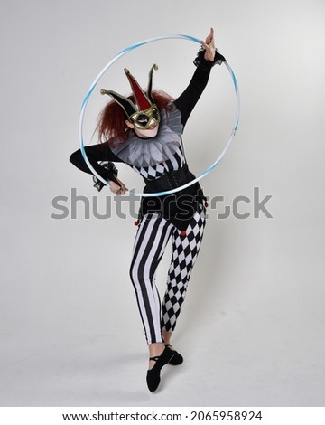 Full length  portrait of red haired  girl wearing a black and white clown jester costume, theatrical circus character.  Standing pose with hula hoop, isolated on  studio background.
