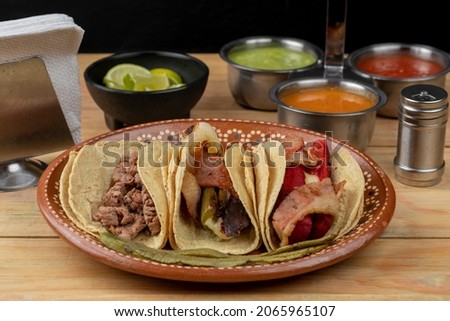 Three tacos served on a clay plate accompanied by salsa and lemons on a wooden table