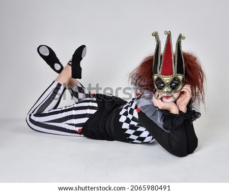 Full length  portrait of red haired  girl wearing a black and white clown jester costume, theatrical circus character.  Sitting down on floor, isolated on  studio background.