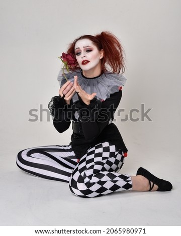Full length  portrait of red haired  girl wearing a black and white clown jester costume, theatrical circus character.  Sitting down on floor, isolated on  studio background.