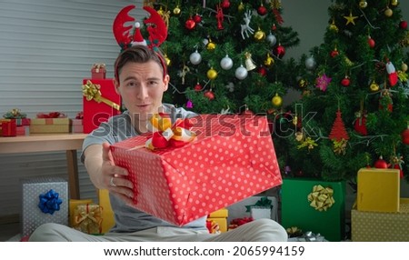 White Caucasian young man, good-looking in concept image, Christmas gift. The young man handed a gift box and looked at the camera. Stack of gift boxes and a Christmas tree decorated in the back.