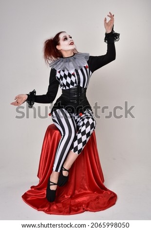 Full length  portrait of red haired  girl wearing a black and white clown jester costume, theatrical circus character.  Sitting down on chair, isolated on  studio background.