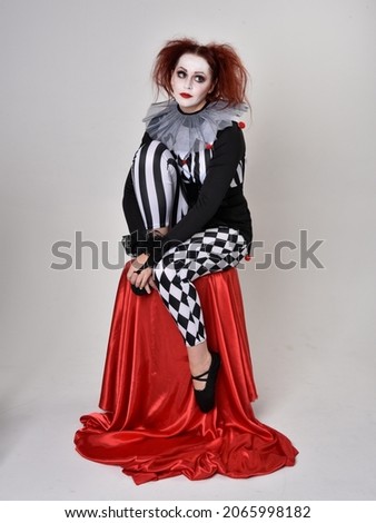 Full length  portrait of red haired  girl wearing a black and white clown jester costume, theatrical circus character.  Sitting down on chair, isolated on  studio background.