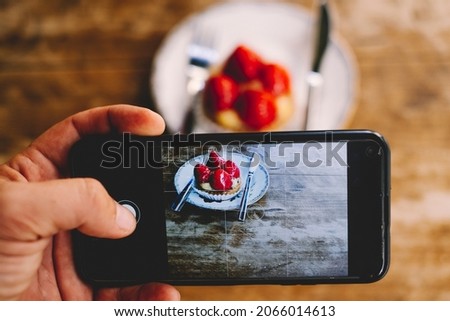 Close up of man taking picture of a little strawberry cake to share on social media and receive likes and followers. People and internet technology. Wooden table