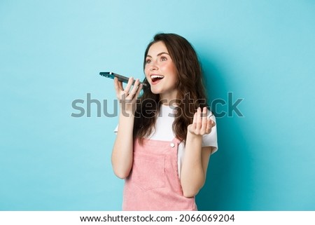 Young woman using app translator on smartphone, speaking into mobile phone dynamic, holding cellphone near lips, recording voice message, blue background