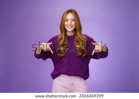 Girl giving recommendation to visit place, pointing down and smiling friendly with delighted and pleasant grin. Portrait of attractive redhead female model in purple warm sweater promoting copy space