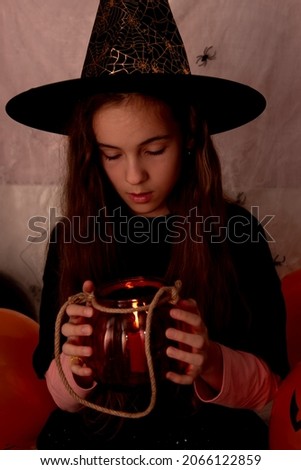 Children's halloween. A smiling girl in a witch hat and an black T-shirt with a glass jar in the shape of a pumpkin is preparing to celebrate. multi-colored balls in the background. Holiday at home.