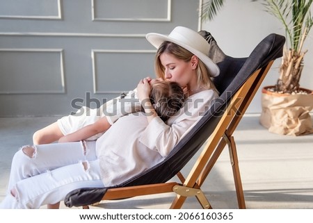 A pregnant mother sits on a wooden deck chair, hugs, kisses her older daughter. A young woman is playing, having fun with a girl. Healthy pregnancy, happy motherhood. Copy space. Loft interior