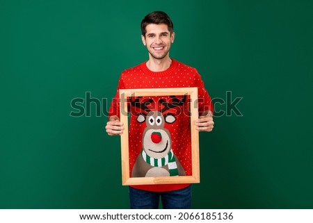 Portrait of attractive cheerful guy holding frame showing deer ornament portrait isolated over green color background