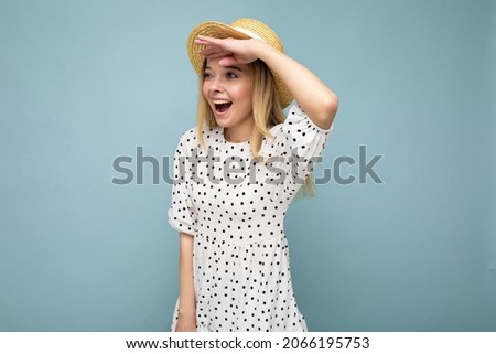 Photo of young positive happy surprised beautiful blonde lady with sincere emotions wearing summer dress and straw hat isolated over blue background with copy space and looking to the side with hand