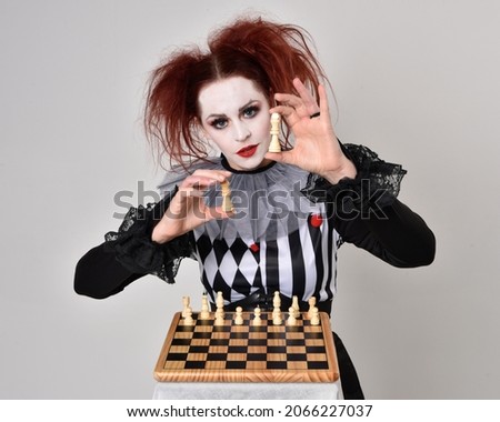 Full length  portrait of red haired  girl wearing a black and white clown jester costume, theatrical circus character.   Playing chess boardgame, isolated on  studio background.