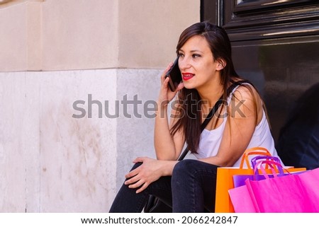 A Beautiful woman sitting on the steps of the entrance of a building with bags on the side and talking on the cell phone smiling..