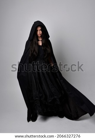 Full length portrait of dark haired girl wearing a witch black flowing gown and  fantasy cloak.   Standing pose with gestural movements, isolated on studio background. Royalty-Free Stock Photo #2066327276