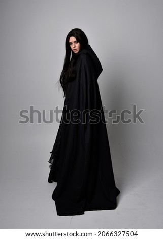 Full length portrait of dark haired girl wearing a witch black flowing gown and  fantasy cloak.   Standing pose with gestural movements, isolated on studio background. Royalty-Free Stock Photo #2066327504