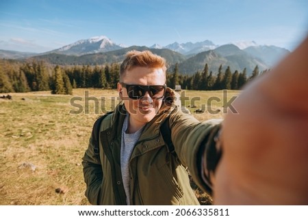 Young man in sunglasses making selfie photo high in the snow mountains enjoying the view. Freedom, happiness, travel and vacations concept, outdoor activities, he wearing a green jacket