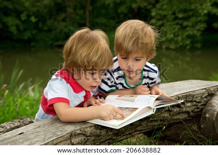 Adorable Little Twin Brothers Looking and Pointing at Very Interesting Picture in the Book Near the Beautiful Lake at Summer