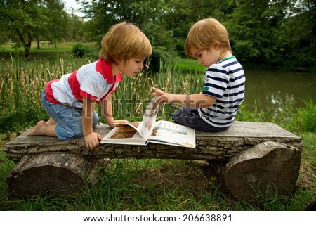 Adorable Little Twin Brothers Sitting on a Wooden Bench and  Looking at Interesting Pictures in the Book Near the Beautiful  Lake at Summer