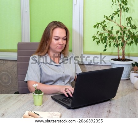 Woman is sitting at table and looking at laptop monitor. There is flowering succulent in pot nearby. Selective focus.