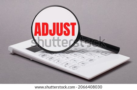 On gray background black calculator and magnifier with text ADJUST. Business concept