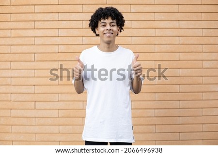 Gesture, emotion and people concept - Smiling African-American guy looking into the camera and seemingly holding a sign on the sides for copy space, dressed in a white t-shirt on a brick wall