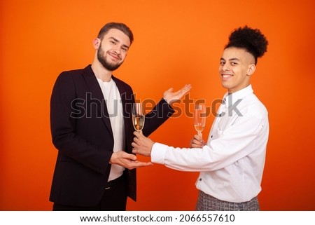 Celebration, holidays, business and people concept - Handsome gay African-American man offering a glass of wine to his bearded male friend at a general corporate party on an orange background