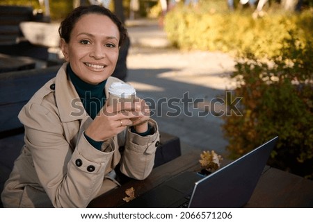 Portrait of a confident beautiful business woman holding a takeaway paper cup with hot drink and smiling toothy smile looking at camera sitting on wooden bench of outdoor cafe in autumn oak grove