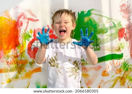 a boy in a white T-shirt, stained with paint, and dirty blue hands, stands against the background of a multi-colored wall. The child painted with his hands. A funny, lovable boy, looks into the frame. Royalty-Free Stock Photo #2066580305