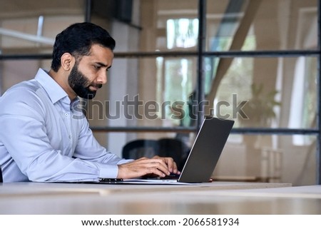 Side view of serious adult concentrated focused Indian Hispanic boss ceo businessman using typing on computer pc laptop working in contemporary office, accounting analysing report financial data. Royalty-Free Stock Photo #2066581934