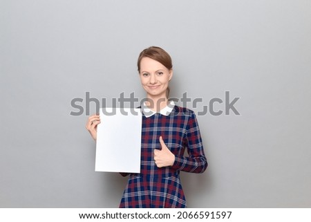 Studio portrait of happy young woman holding white blank paper sheet with place for your text in hand, raising thumb up in approval and like gesture, smiling cheerfully, standing over gray background